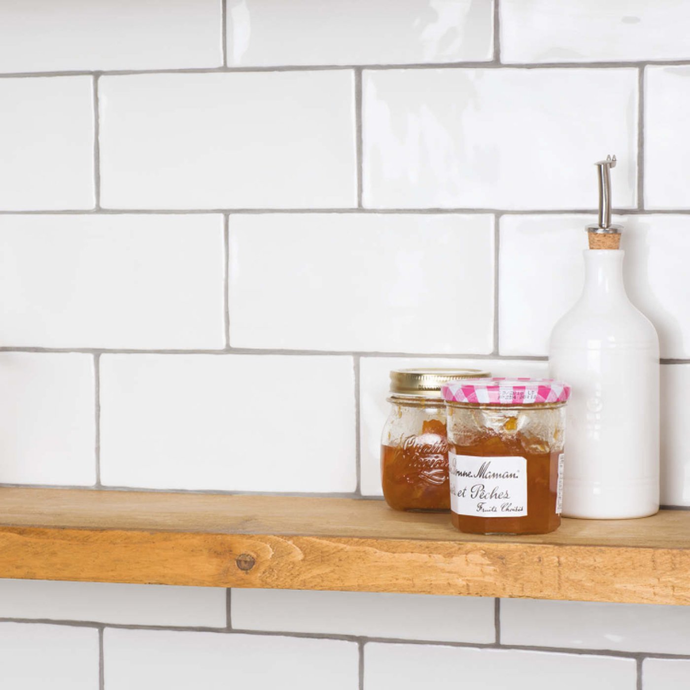 Image of A rustic wooden floating shelf mounted against a white subway tile wall, holding two glass jars of jam and a white ceramic oil bottle with a metal pourer.