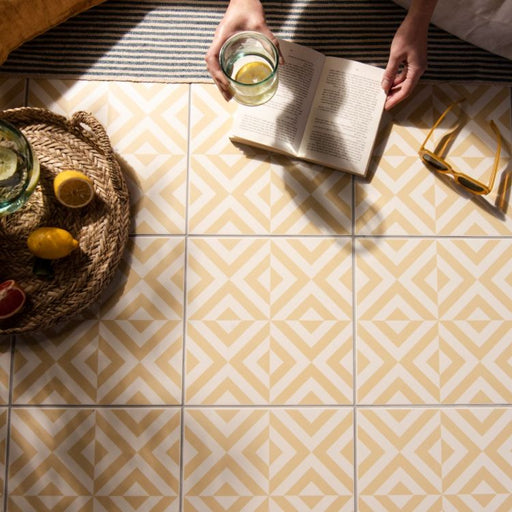 yellow and white geometric floor tiles with an open book, sunglasses, and a glass of water with lemon on the floor beside a woven tray of fruit.