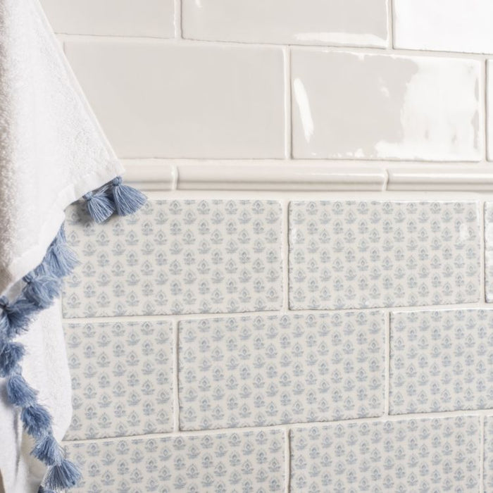 Close-up of a tiled wall with glossy white tiles on top and patterned blue-and-white tiles below, with a white towel featuring blue tassels hanging on the left.