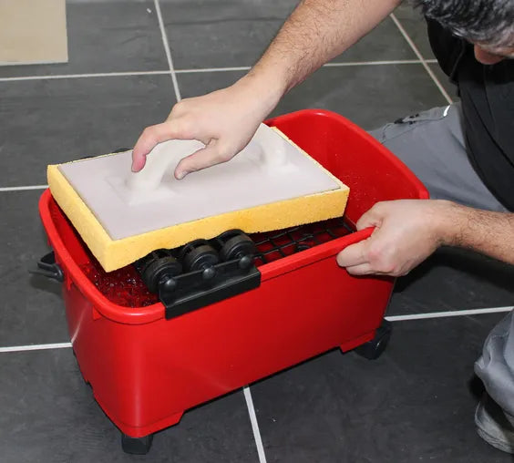 Image of Person using a red cleaning bucket with sponges and a scrubber on a tiled floor.