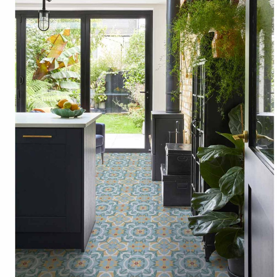 Cabana Sicily Patterned tiles 20x20cm as kitchen floor tiles. Dark kitchen cabinets underneath white worktops. Leading to black framed bi-fold doors, log burner and large, green plants. Bookcase and box storage to the walls.
