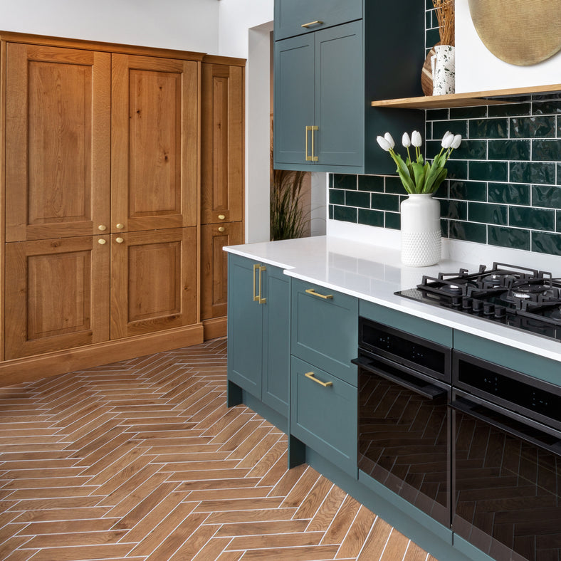 Elegant Parquet Wood effect tiles 8x33cm in a kitchen setting.  Teal kitchen cabinets with a white worktop, green metro tiles on the wall as splashback tiles. cast iron hob with white vase and flowers next to it. Cupboard with wooden doors to the rear. Farmhouse kitchen replicating cottagecore.