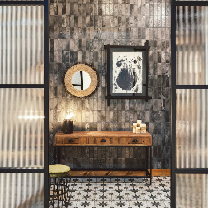 Raku Black wall tile 20x40cm in a hallway setting with a wooden console table. A mirror sits on the tiled wall, with a black and white picture of a vase. Candles sit on the console table while white screens frame either side of the tiles.
