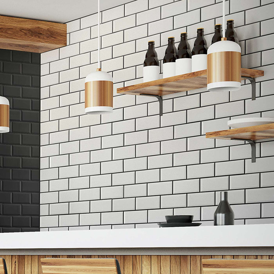 Metro Matt White Bevelled Brick tile 10x20cm in a kitchen setting. White brick tiles with black grout on the kitchen wall with white worktops, wooden kitchen cabinets. Wooden shelves on the wall with dark glass bottles on them.